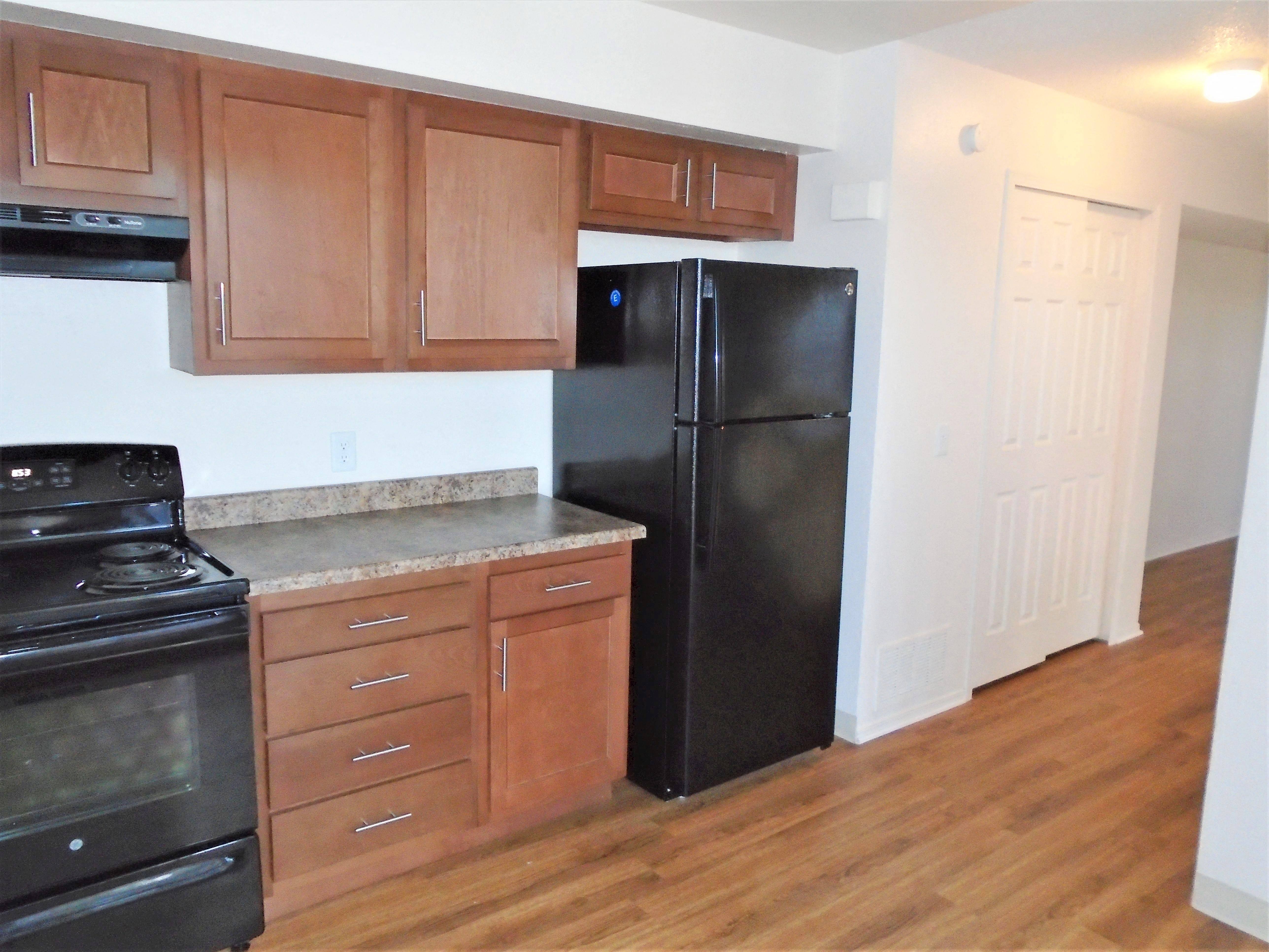 A kitchen with light brown counter tops, brown cabinets, stove, refrigerator, pantry with sliding door, wood like flooring, and white walls.